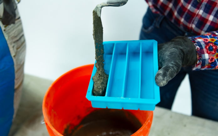 Una mano enguantada utiliza una paleta de albañilería para verter la mezcla de hormigón en una bandeja de hielo.
