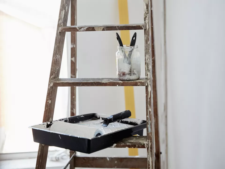 Una escalera de madera salpicada de pintura, un frasco con dos pinceles y una bandeja de pintura y un rodillo.